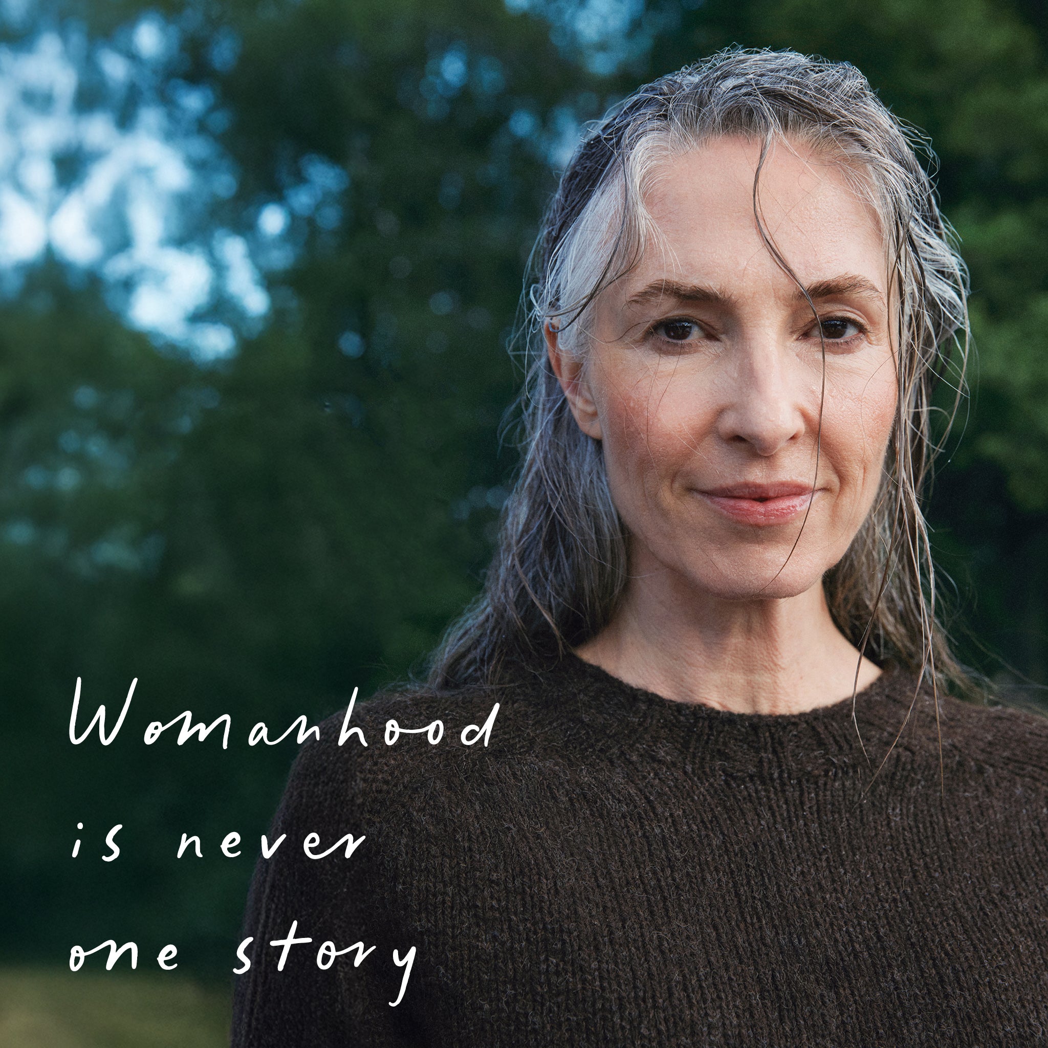 Empowering campaign image of woman with grey hair and quote ‘Womanhood is never one story’ on blurred natural background