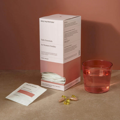 Daily Essentials for Women's Fertility supplements with sachets, pills, and a glass of water on a stone surface.