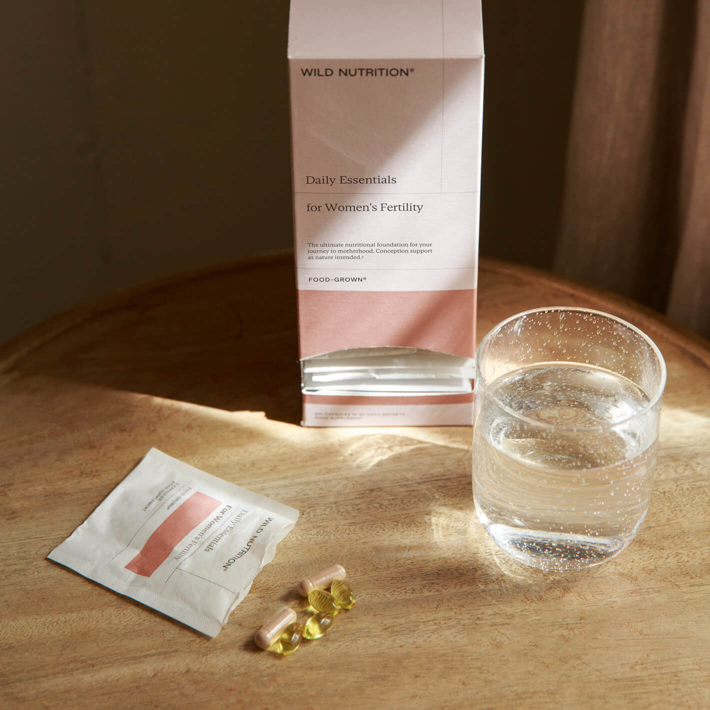 Daily Essentials for Women's Fertility supplements with water on a wooden table, featuring omega-3 and prenatal support.