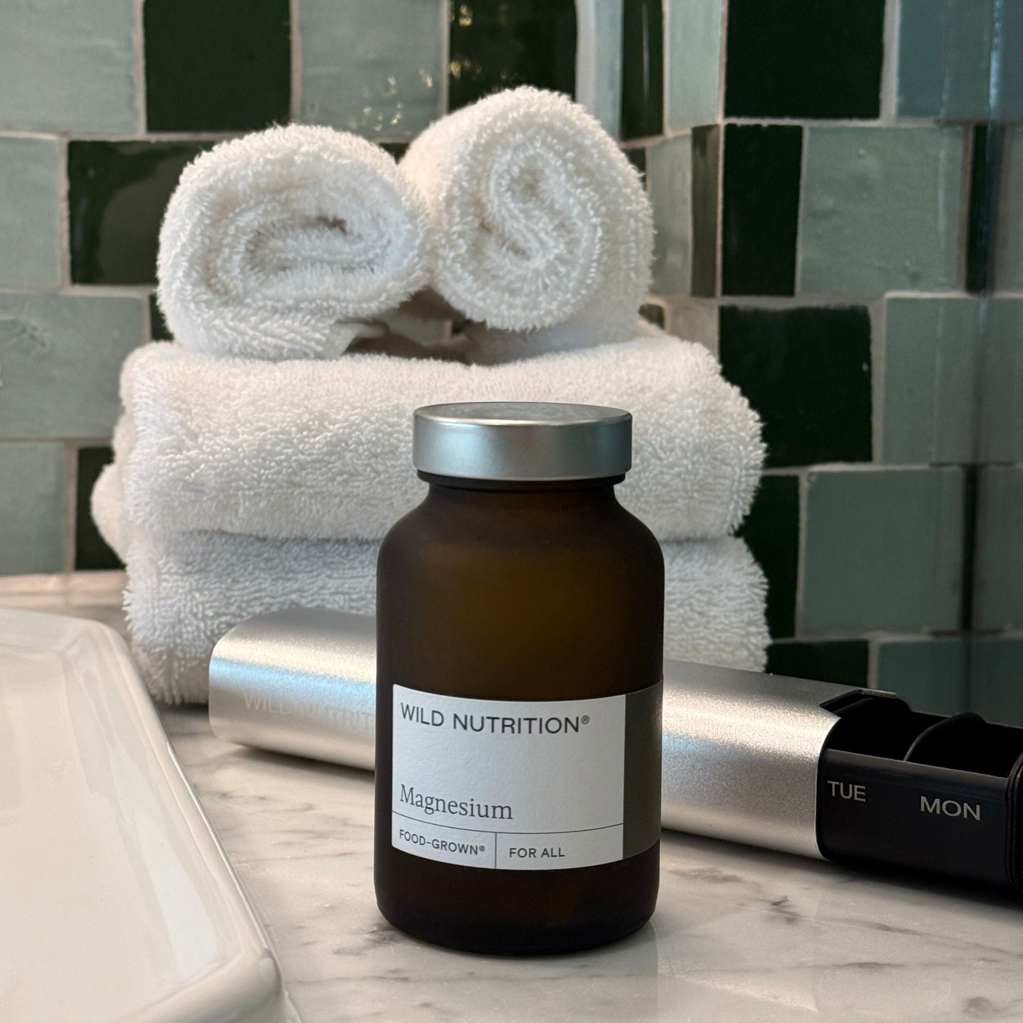 Wild Nutrition Food-Grown Magnesium supplement bottle on marble bathroom counter with capsule dispenser and rolled white towels