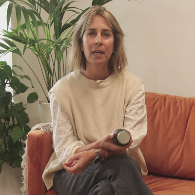 Woman in a beige sweater holding a bottle while sitting on an orange couch surrounded by plants.