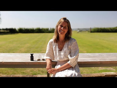 Smiling woman in a white dress sitting at a wooden table outdoors with Food-Grown® Natural Glow supplement.